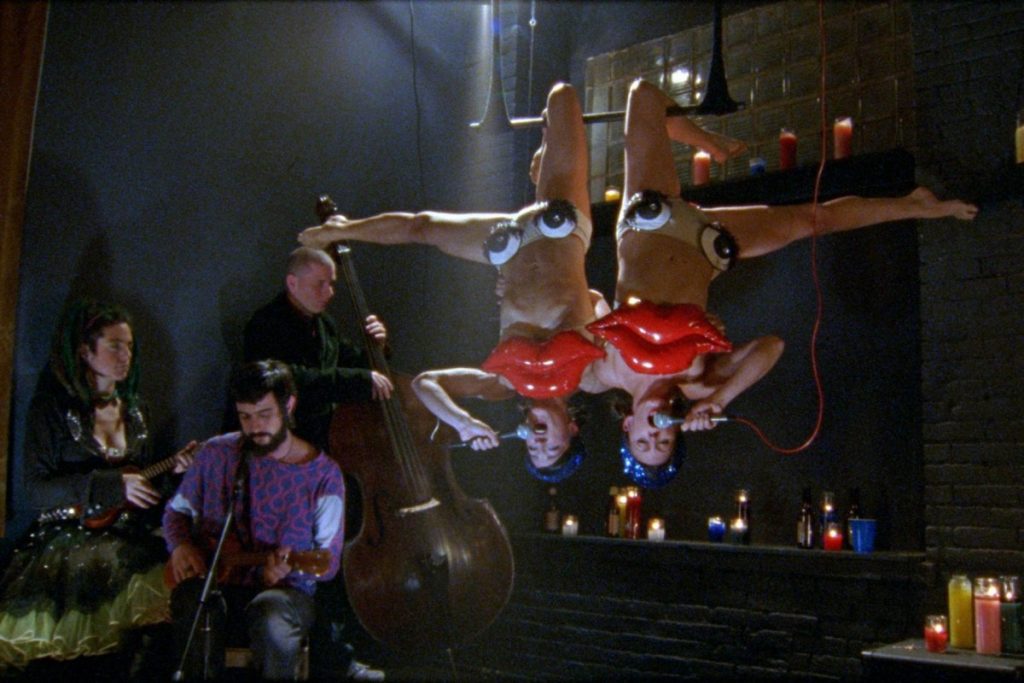 A scene showcasing the inside of the sex club Shortbus. A man plays the guitar on the right side and two female performers in red and white dresses hang upside down at the center of the image. They wear big googly eyes.
